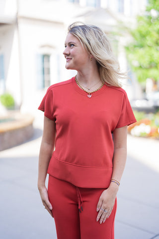 A close-up portrait view of the SPANX Crewneck Tee highlighting the ultra-smooth SPANX AirEssentials® spacer fabric texture in a bold red shade. Details show the ribbed crew collar, clean shoulder stitching, and wide hem band. Styled with layered gold necklaces, gold hoop earrings, and delicate hand jewelry.