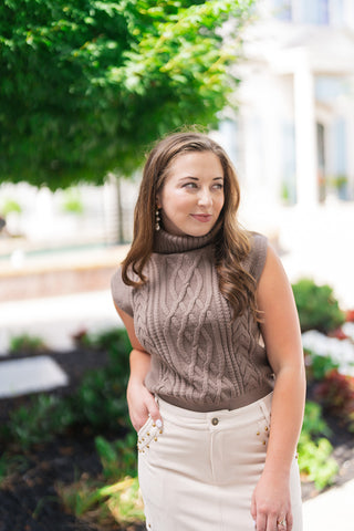 A portrait view of the Driftwood Braided Cable Top highlighting the rich, intricate texture of the braided cable stitches running down the center bodice. The shot details the cozy fold-over turtleneck, structured cap sleeves, and the wide ribbed hem tucked neatly into the waistband of a high-waisted cream skirt.