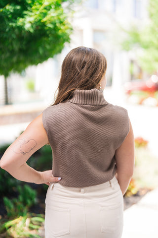 Back view of the Driftwood Braided Cable Top demonstrating the textured, waffle-weave knit construction across the back and shoulders. The image showcases the ribbed fold-over turtleneck collar, full armhole coverage along the cap sleeves, and the clean fit resting over a high-waisted cream mini skirt.