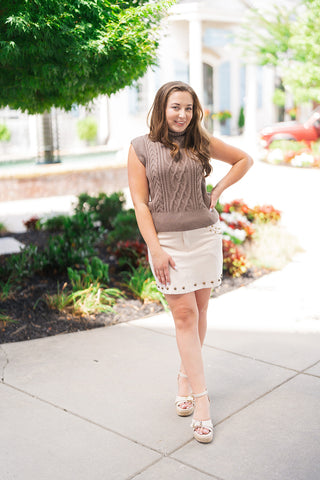 Woman posing outdoors in the Driftwood Braided Cable Top—a taupe-brown sleeveless sweater top featuring a thick fold-over turtleneck collar, center braided cable-knit panels, and cap-sleeve arm openings. Styled for a chic transitional look paired with a cream studded A-line mini skirt, off-white platform espadrille sandals, and drop earrings.