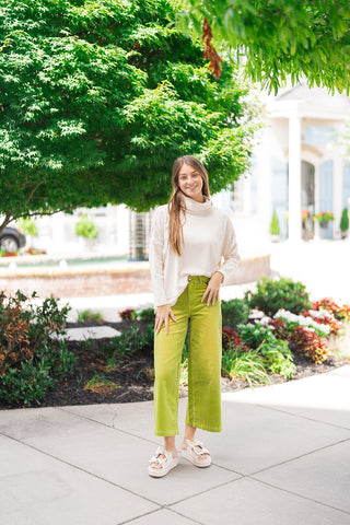 Full-length view of a smiling woman standing outdoors in the Vanilla Cloud Relaxed Knit—an ultra-soft, cream-vanilla long-sleeve sweater featuring a draped cowl turtleneck, drop-shoulder seams, and a relaxed boxy fit. Styled effortlessly with chartreuse green high-waisted corduroy pants and off-white bow platform slide sandals.