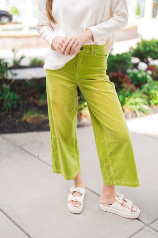 A close-up view highlighting the front details of the high-waisted Matcha Latte Corduroy Pants. The rich, chartreuse green fine-wale corduroy features a classic zip-fly with a single button closure, belt loops, and traditional front pockets, flowing effortlessly into a relaxed, wide-leg cropped silhouette that rests right above the ankle.