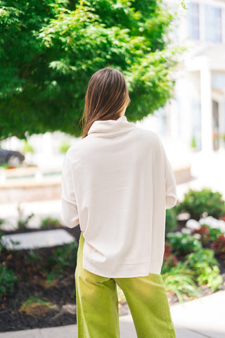 Back view of the Vanilla Cloud Relaxed Knit demonstrating the clean, minimal drape across the back and shoulders. The shot emphasizes the relaxed posture of the draped cowl collar, the relaxed boxy silhouette, and the extra length provided by the subtle high-low stepped hemline.