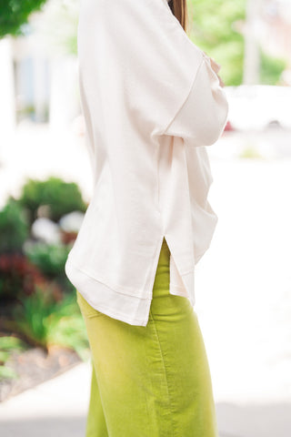 A side-profile close-up view focusing on the lower construction and fabric drape of the Vanilla Cloud Relaxed Knit. The shot details the modern high-low hemline with a high side vent, exposed topstitching along the seam, and the loose dolman-style sleeve resting over chartreuse corduroy pants.