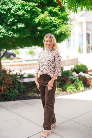 A full-length view of a smiling blonde woman posing outdoors in the Blush Leopard Mock Neck Sweater—a soft blush pink pullover knit featuring an allover leopard print in neutral olive tones, a sophisticated mock turtleneck collar, and wide 3/4-length drop-shoulder sleeves. She has styled the top with high-waisted dark brown wide-leg trousers, woven neutral block heel sandals, and a gold chain choker necklace.