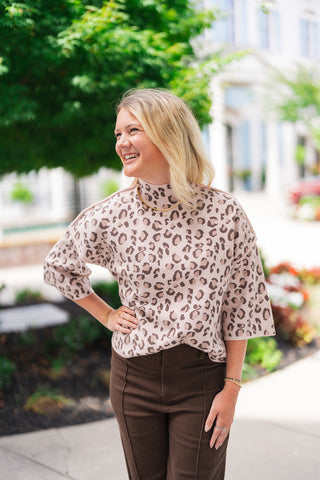 A portrait close-up view highlighting the front construction of the Blush Leopard Mock Neck Sweater. The image details the cozy mock turtleneck collar, the gold chain necklace resting over the knit, the prominent drop-shoulder seam detailing, and the fine-gauge texture of the soft blush pink and olive animal print fabric.