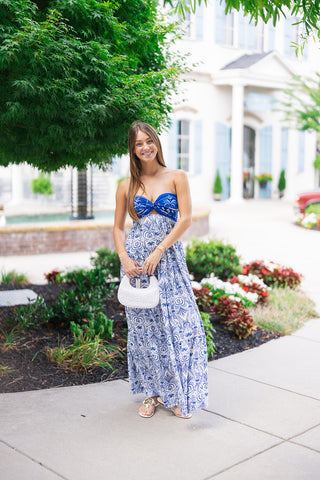 Woman posing in the Cobalt Tile & Twist Maxi Dress, a strapless vacation dress featuring a rich cobalt blue twisted bust with a subtle keyhole cutout. The flowing, tiered maxi skirt showcases an intricate navy and blue Mediterranean tile and botanical pattern over a light backdrop. Styled for resort wear with a structured white leather top-handle purse and white flat thong sandals in front of a coastal resort setting.