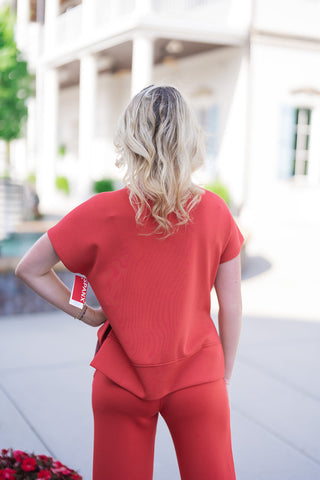 Back view of the SPANX Crewneck Tee in vibrant red, showcasing the relaxed silhouette, dropped shoulder construction, and high-low hem design that provides extended back coverage. The model stands turned away with one hand on her hip, showing the slight side-vent opening over matching red pants.