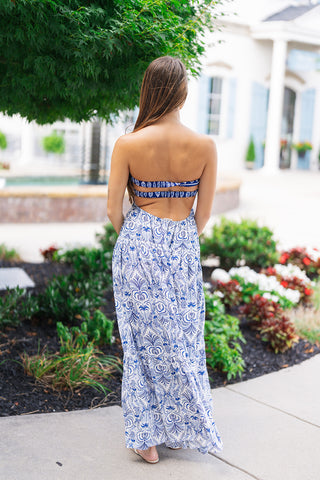 Back view of the Cobalt Tile & Twist Maxi Dress displaying the open-back design anchored by double smocked cobalt blue elastic bands for a flexible, stay-put fit. The flowing tiered maxi skirt cascades down to ankle length, showcasing the full tilework print as the model stands outdoors near lush greenery and stone architecture.