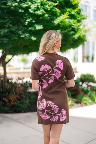 Back view of the Woodland Whimsy Sweater Dress showcasing the continuous light pink ginkgo-leaf branch pattern flowing from the shoulder across the lower back and hemline. The model is turned away, highlighting the straight-cut mini silhouette, short sleeves, and fine knit texture of the chocolate brown fabric.