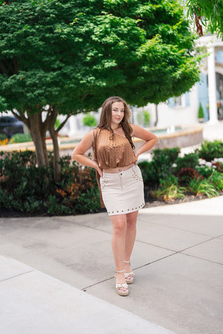 Full-length front view of a woman posing outdoors in the Sunset Soirée One-Shoulder Top—a bronze camel satin blouse featuring sharp vertical accordion pleats, an asymmetrical single-shoulder neckline, and a voluminous elastic bubble hem. The top features a long, dramatic fabric panel cascading down from the shoulder strap behind her back. She has styled it with a high-waisted off-white studded denim mini skirt and platform ankle-strap wedges.
