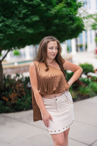Close-up view of the Sunset Soirée One-Shoulder Top highlighting the rich texture and luminous sheen of the camel bronze pleated satin. The detail shot emphasizes the dramatic vertical pleats, the softly gathered bubble hemline at the waist, and the asymmetrical neckline framing the collarbone.