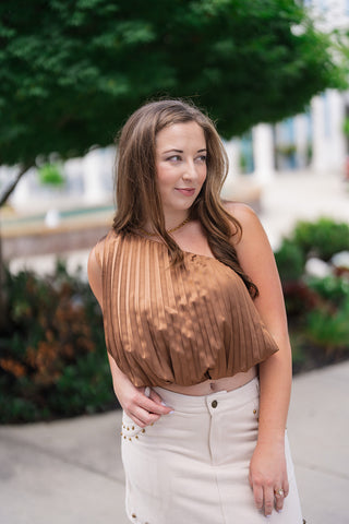A mid-length portrait shot highlighting the front fit and structure of the Sunset Soirée One-Shoulder Top. The image details the fine accordion pleating across the silky woven satin fabric, the cropped gathered bubble waist, and the single-shoulder neckline paired with a gold necklace and an off-white studded skirt.