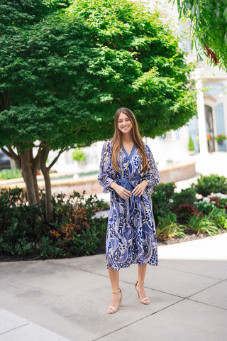 Woman smiling in the Sapphire Serenade Midi Dress, a sweeping midi-length dress featuring a vibrant abstract botanical and animal spot print in sapphire blue, periwinkle, cream, and black tones. Designed with a deep V-neckline framed by a blue and white gingham check trim and a matching gingham self-tie belt at the waist. Styled with nude ankle-strap block heel sandals outdoors in front of a lush green tree and resort architecture.