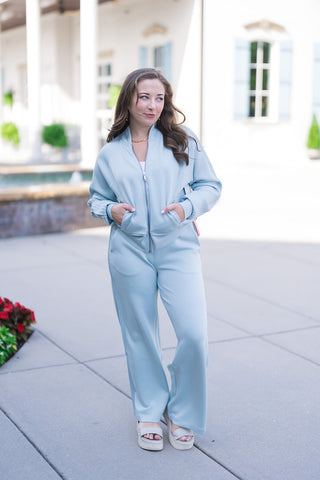 Woman posing in the SPANX Pintuck Track Jacket, an ice-blue full-zip activewear jacket made from SPANX AirEssentials® spacer fabric. Styled as a monochromatic athleisure set with matching ice-blue wide-leg pants and off-white platform slide sandals. The jacket features a silver zip closure, sporty mockneck collar, raglan sleeves with pin-tuck stripe accents along the arms, and convenient front pockets where the model rests her hands.