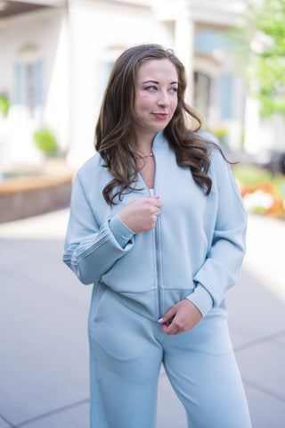 A mid-shot portrait view highlighting the details of the ice-blue SPANX Pintuck Track Jacket. The close-up showcases the smooth AirEssentials® spacer fabric, silver front zipper, relaxed raglan shoulder construction, and wide ribbed sleeve cuffs. The model is looking to the side with dark brown wavy hair, styled with a subtle gold necklace.