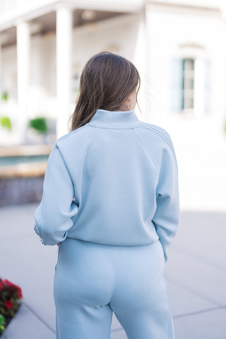 Back angle close-up of the SPANX Pintuck Track Jacket in soft ice-blue. Shows the back of the structured mockneck collar, clean back yoke construction, raglan shoulder seams, and the raised pintuck stripe detailing running down the sleeves. Styled with matching ice-blue bottoms in an outdoor architectural setting.