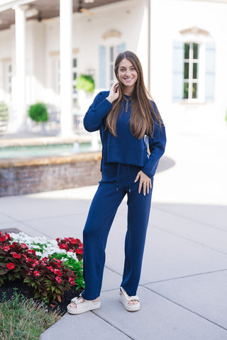 Woman smiling in the SPANX Crewneck Long Sleeve in solid navy blue, paired with matching navy drawstring lounge pants for a set look. The relaxed-fit pullover top features dropped shoulders, a classic crew neckline, and a subtle side-split hem. Styled with off-white platform slide sandals and a gold Y-necklace on a paved outdoor courtyard path near white and red flowerbeds
