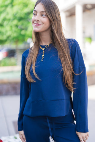 A close-up portrait view of the SPANX Crewneck Long Sleeve highlighting the ultra-smooth SPANX AirEssentials® spacer fabric texture and classic crew neckline. Details show the relaxed fit, dropped shoulder seams, subtle side vents, and a wide stitched bottom hem. Accessorized with long straight brown hair and a gold chain necklace.