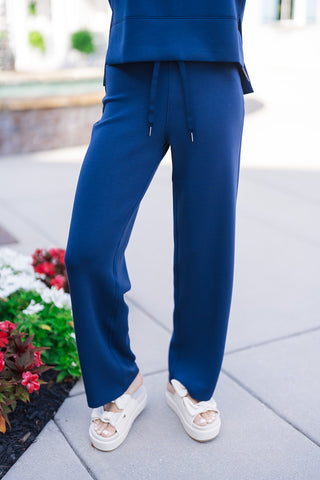 A close-up view of the SPANX Straight Leg Pants in navy blue, focusing on the high-waisted fit, tonal drawstring ties with metallic aglet tips, and smooth front seams. The straight legs drop cleanly to the ankles over off-white platform sandals near red and white flowerbeds.