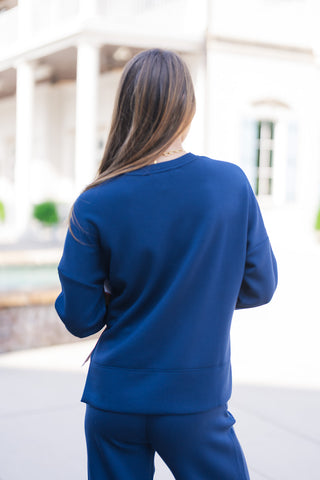 Back angle view of the SPANX Crewneck Long Sleeve in deep navy blue, displaying the clean back yoke construction, dropped shoulders, and straight hem line that provides generous back coverage. The model is turned away, wearing matching navy pants in an outdoor setting with light-colored European-style architecture in the background.