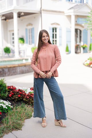 Woman posing outdoors in the Blush & Peplum Cable Pullover—a dusty rose pink mixed-media knit top featuring radiating chest cable-knit lines, a ribbed mock turtleneck, long cable-knit sleeves, and a matching woven peplum ruffle hem. Styled for a chic, casual fall outfit paired with wide-leg blue denim jeans, neutral straw platform wedges, and a gold cross pendant necklace in front of a white brick building.