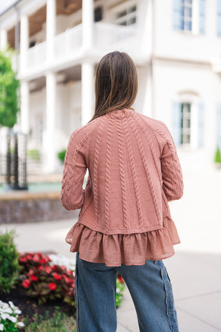 Back view of the Blush & Peplum Cable Pullover showcasing the fan-like, radiating cable-knit pattern spreading across the upper back and shoulders. The shot emphasizes the full coverage of the long cable sleeves, the seamless flow into the gathered woven peplum ruffle hem, and the relaxed fit paired with wide-leg jeans.