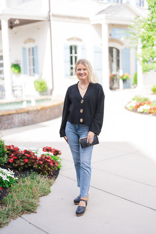 A smiling blonde woman posing outdoors in the Polished Gold & Onyx Cardigan—a soft, fine-gauge onyx black V-neck knit sweater cardigan adorned with three oversized, high-shine polished gold statement buttons down the front. Styled for an elevated casual look with light-wash straight-leg denim jeans, black wooden clog heels, a gold chain necklace, and a studded black leather clutch handbag.