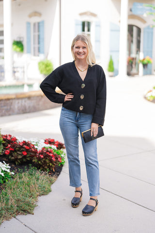 Full-length front view of a blonde woman standing outdoors in the Judy Blue Peggy Ankle Straight Jeans—high-waisted medium-wash indigo blue denim pants featuring subtle whiskering, authentic thigh fading, and a straight, ankle-length cut. Styled for an elevated casual look paired with a black V-neck cardigan sweater with gold statement buttons, black studded wooden clog heels, and a gold chain necklace.