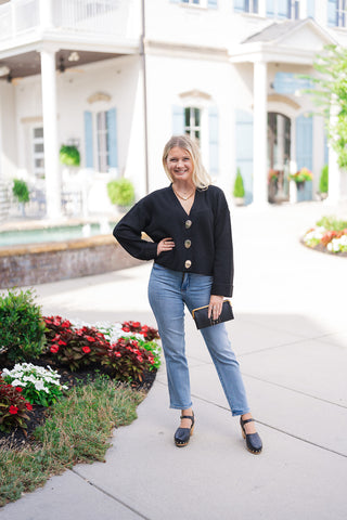 Full-length view of a woman posing with one hand on her hip wearing the Polished Gold & Onyx Cardigan. The shot highlights the relaxed, slightly cropped silhouette, drop-shoulder seams, and wide ribbed hemline sitting at the waist of high-waisted jeans, framed by a boutique building background.