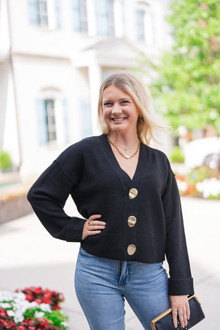 A portrait close-up view highlighting the front construction of the Polished Gold & Onyx Cardigan. The detail shot emphasizes the deep V-neckline framing the collarbone, the high-end metallic shine of the oversized gold buttons, and the soft texture of the fine-gauge black knit fabric.