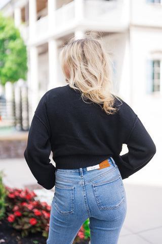 Back view of the Polished Gold & Onyx Cardigan demonstrating the clean, smooth knit texture across the shoulders and back. The view showcases the drop-shoulder sleeve seams and the thick ribbed waist hem resting above high-rise light-wash denim jeans with back patch pockets.