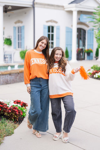 Two women posing together in the Tennessee Cloudy Ringer Sweatshirt, showcasing both colorways. The model on the left wears the vibrant orange cloudy knit pullover with white trim paired with wide-leg blue jeans. The model on the right wears the cream-colored cloudy knit pullover with orange trim paired with washed charcoal jeans, holding an orange game day pom-pom outdoors.