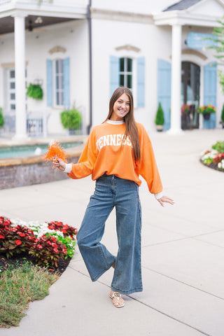 Woman posing in the orange colorway of the Tennessee Cloudy Ringer Sweatshirt, featuring white arched "TENNESSEE" block lettering and contrasting white ringer trim along the collar, cuffs, and hemline. Styled with wide-leg blue jeans and white flat sandals while holding an orange pom-pom.