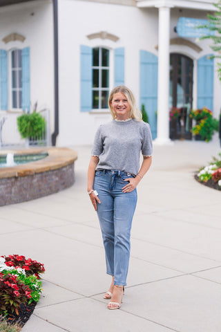 Woman smiling in the Heathered Honey Scallop Knit Top, a short-sleeve sweater in a versatile heather grey knit. The top features subtle puff shoulders, a fitted ribbed hem, and delicate cream-colored scalloped lace trim along the crew neckline and sleeve cuffs. Styled for an everyday transitional look with high-waisted light-wash straight-leg jeans, nude ankle-strap heels, and subtle gold jewelry on a paved outdoor walkway.