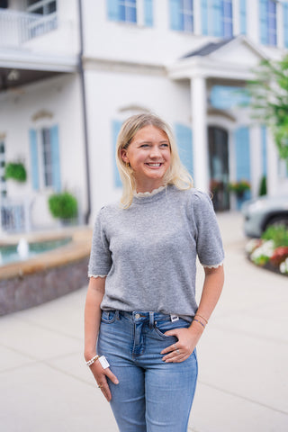 A portrait view of the Heathered Honey Scallop Knit Top highlighting the intricate cream scalloped trim detailing along the ribbed crew collar and short puff sleeve cuffs. The cozy, soft-textured heather grey knit fits smoothly across the bodice, tucked neatly into light blue denim jeans with stacked bead bracelets.