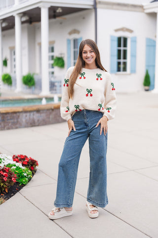 Full-length front view of a smiling woman posing outdoors in the Cherry on Top Fuzzy Knit—a cozy, plush eyelash knit sweater in a cream shade adorned with 3D red cherry pom-poms and green yarn bow motifs across the front and long raglan sleeves. Styled casually with high-waisted medium-wash wide-leg denim jeans and off-white platform bow slides.
