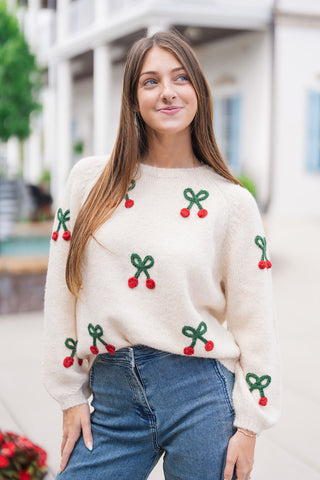A portrait close-up view highlighting the detailed texture of the Cherry on Top Fuzzy Knit. The image showcases the soft, fuzzy cream yarn, the classic ribbed crew neck, raglan shoulder seams, and the raised, tactile 3D cherry pom-poms with tied green yarn bows spread across the chest and long sleeves.