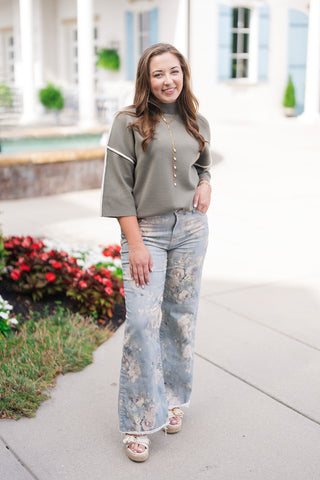 A full-length front view of a woman posing outdoors in the Olive Grove Contrast Sweater—a soft, fine-gauge mock neck sweater top in a sage olive green hue featuring crisp white piping along the drop shoulders and 3/4-length sleeves. She has styled the relaxed, boxy knit with high-waisted floral print wide-leg jeans, off-white platform espadrille sandals, and a long gold pearl lariat necklace.