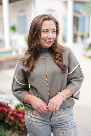 A portrait close-up view highlighting the front details of the Olive Grove Contrast Sweater. The image showcases the sleek mock turtleneck collar, fine-gauge smooth knit texture, and the contrasting white piping that outlines the relaxed drop-shoulder seams and 3/4 sleeves, accessorized with a gold chain lariat necklace.