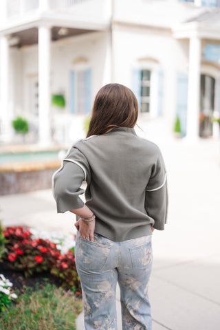 Back view of the Olive Grove Contrast Sweater demonstrating the clean, minimalist look across the back. The shot highlights the continuation of the white piping accent running over the top of the drop shoulders down the wide 3/4 sleeves, as well as the straight hemline resting over high-rise floral denim pants.