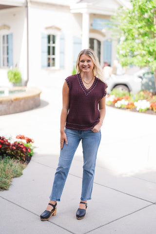 A full-length front view of a smiling blonde woman posing outdoors in the Dark Cocoa Stitch-Trim Sweater Vest—a deep cocoa-brown cable-knit sleeveless top featuring light powder-blue pick-stitching along the V-neckline, cap-sleeve arm openings, and straight hemline. She has styled the sweater vest as a standalone top paired with high-waisted light-wash straight-leg jeans, black leather wooden clog heels, and a gold heart pendant necklace.