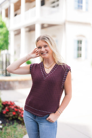 A mid-length front-facing shot highlighting the relaxed drape and texture of the Dark Cocoa Stitch-Trim Sweater Vest. The view details the rich cable-knit weave throughout the body, the contrasting light blue whipstitch trim tracing the V-neck and cap sleeves, and the straight hem with double-line stitch details worn over light-wash denim.