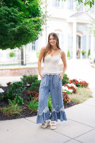 A full-length view of a smiling woman standing outdoors wearing the Flirty Flare Ruffle Denim Pants—light-to-mid-wash chambray denim pants featuring a wide-leg silhouette, gathered drawstring waistband, vertical raw-edge frayed seam accents, and tonal diamond patchwork with tiered ruffle flounce hems. She has styled them with a white lace-trimmed camisole top, off-white bow platform sandals, and gold pendant necklaces.