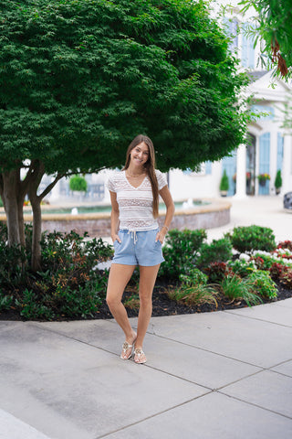 A full-length front view of a woman smiling and posing in the White Willow Lace Blouse. This romantic, bohemian white top features a delicate trimmed V-neckline and horizontal tiers of intricate geometric floral lace panels. She styles the semi-sheer blouse with light blue drawstring shorts and white t-strap slide sandals while standing on a concrete walkway outdoors.