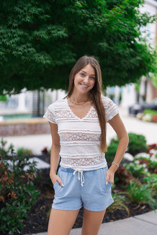 A mid-length front view showcasing the comfortable regular fit of the White Willow Lace Blouse. The design highlights alternating panels of detailed geometric lace and soft, crinkled woven bands. The model stands with her hands in the pockets of light blue shorts, emphasizing the blouse's layered tiered short sleeves with raw edge detailing.