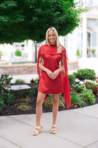 A full-length front view of a blonde woman smiling and posing in the Crimson Crush Scarf Dress. The mini dress features a solid deep red colorway in a premium satin fabric. In this image, the attached crimson chiffon scarf is draped forward across her shoulders and chest like a lightweight poncho, layered over the satin body. She styles the outfit with neutral woven two-strap block-heeled sandals while standing outdoors on a sidewalk.