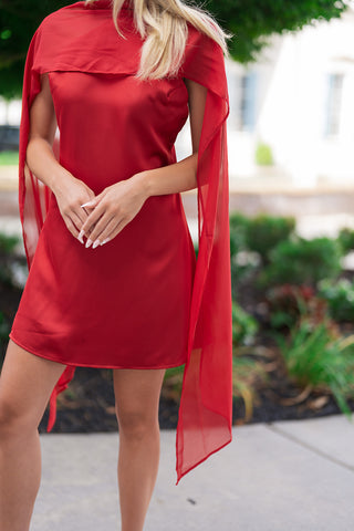 An upper-body portrait view of the Crimson Crush Scarf Dress, showcasing the rich texture contrast between the fluid red satin body and the sheer chiffon scarf. The scarf panel is styled forward, draping softly over her chest and arms to add a romantic layer of multi-dimensional texture. Her hands are elegantly clasped at the waist, showing off white manicured nails.