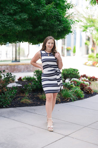 A full-length front view of a woman smiling and posing in the Z Supply Mattie Striped Mini Dress. This sleeveless knit jersey dress features a crisp white and washed ash black horizontal striped pattern. The silhouette is detailed with a structured high neck crewline and a flattering fabric tie accent wrapped around the waist. She is styled with beige platform wedge sandals, a gold pendant coin necklace, and delicate beaded bracelets.