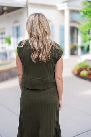 Back view highlighting the textural details and comfortable fit of the olive green ribbed knit set. The image showcases the cap-sleeve top's structured design, contrasting the vertical ribbing of the main bodice with a wide, horizontally ribbed bottom band. The top rests cleanly over the waistband of the matching vertically ribbed midi skirt, creating a seamless and sophisticated silhouette.